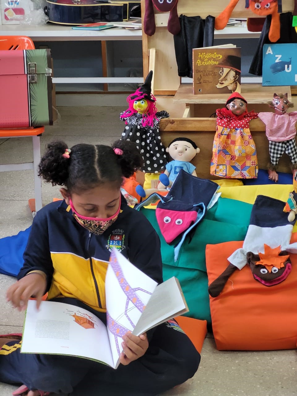 Menina sentada no chão da escola entre as almofadas, lendo o livro que segura com suas mãos e apoia em seu colo.