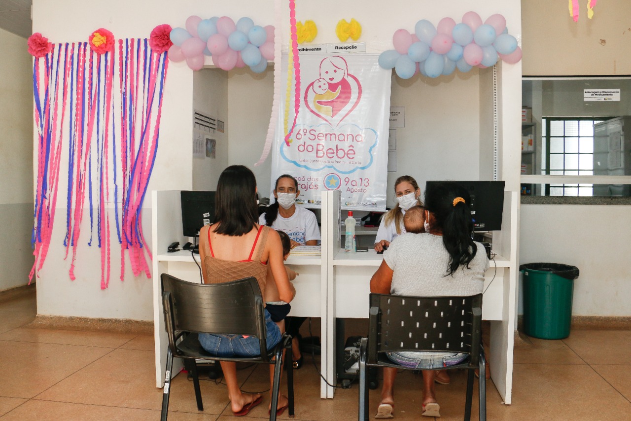 Imagem de quatro mulheres sentadas nas suas respectivas cadeiras: duas mães segurando as crianças no colo de frente a outras duas mulheres com a camisa do evento “Semana do bebê”.