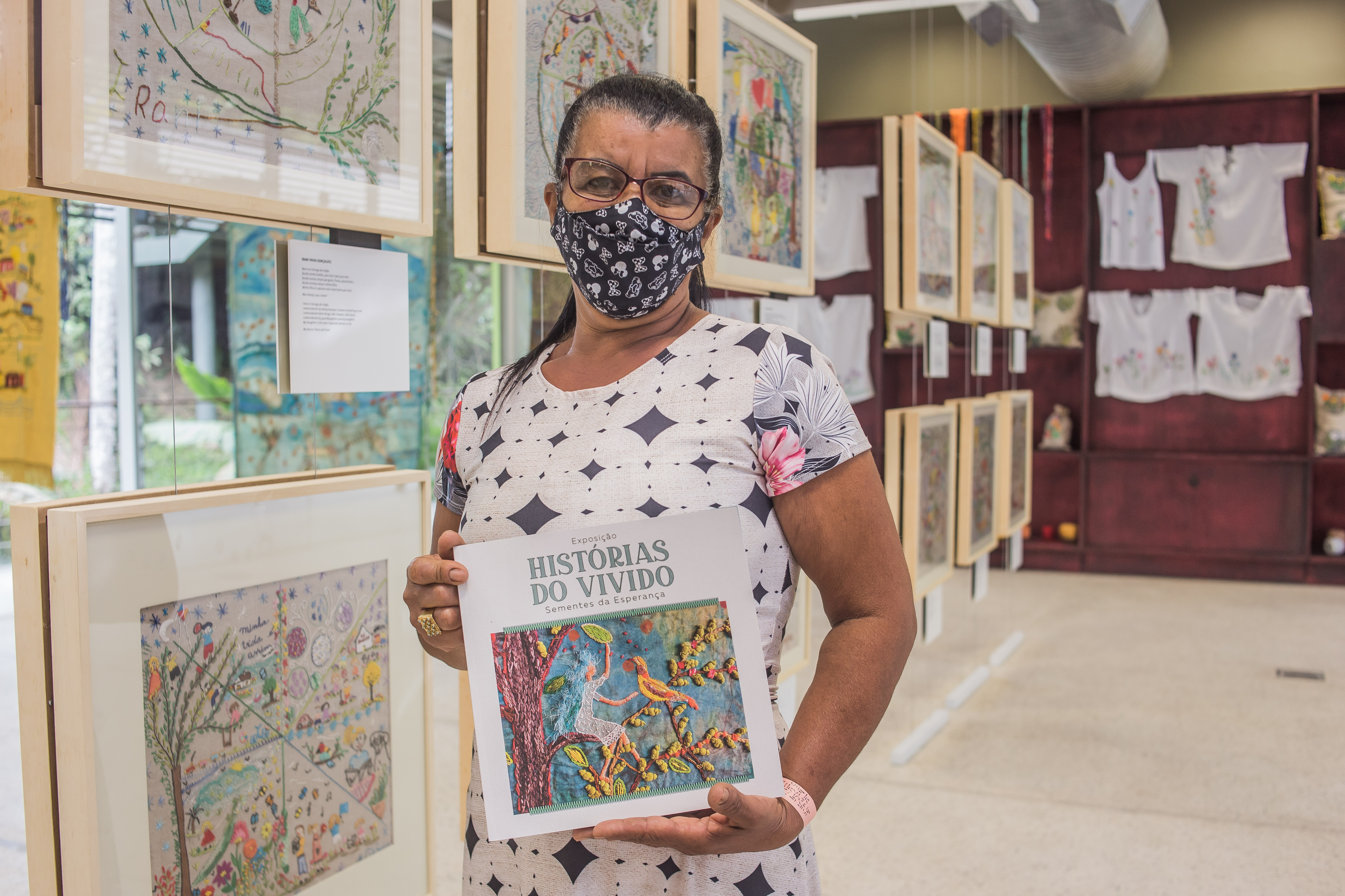 Foto de uma senhora de óculos, máscara e cabelo preso para trás em frente às obras de bordados expostas segurando o guia da exposição “Histórias do vivido” do projeto Semeando Esperança.