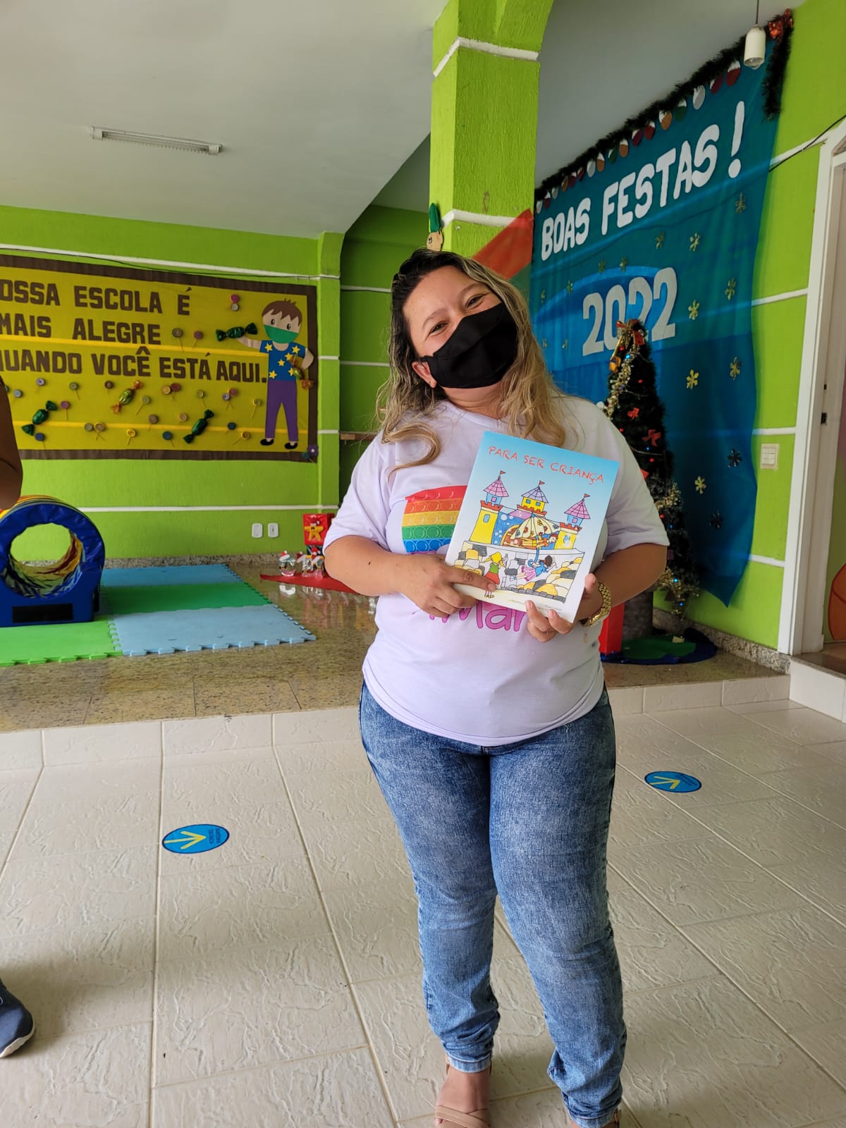 Uma mulher de cabelos claros usando máscara segurando o livro infantil que recebeu “Para ser criança”
