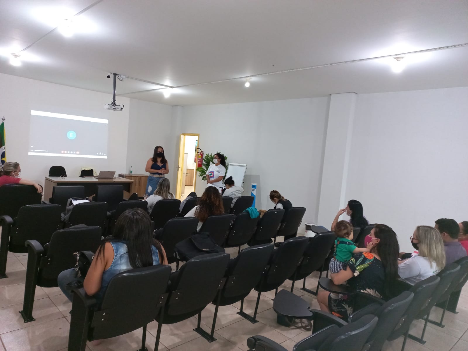 Imagens de pessoas sentadas em cadeiras em uma sala de frente a um telão para a primeira reunião do comitê intersetorial. 