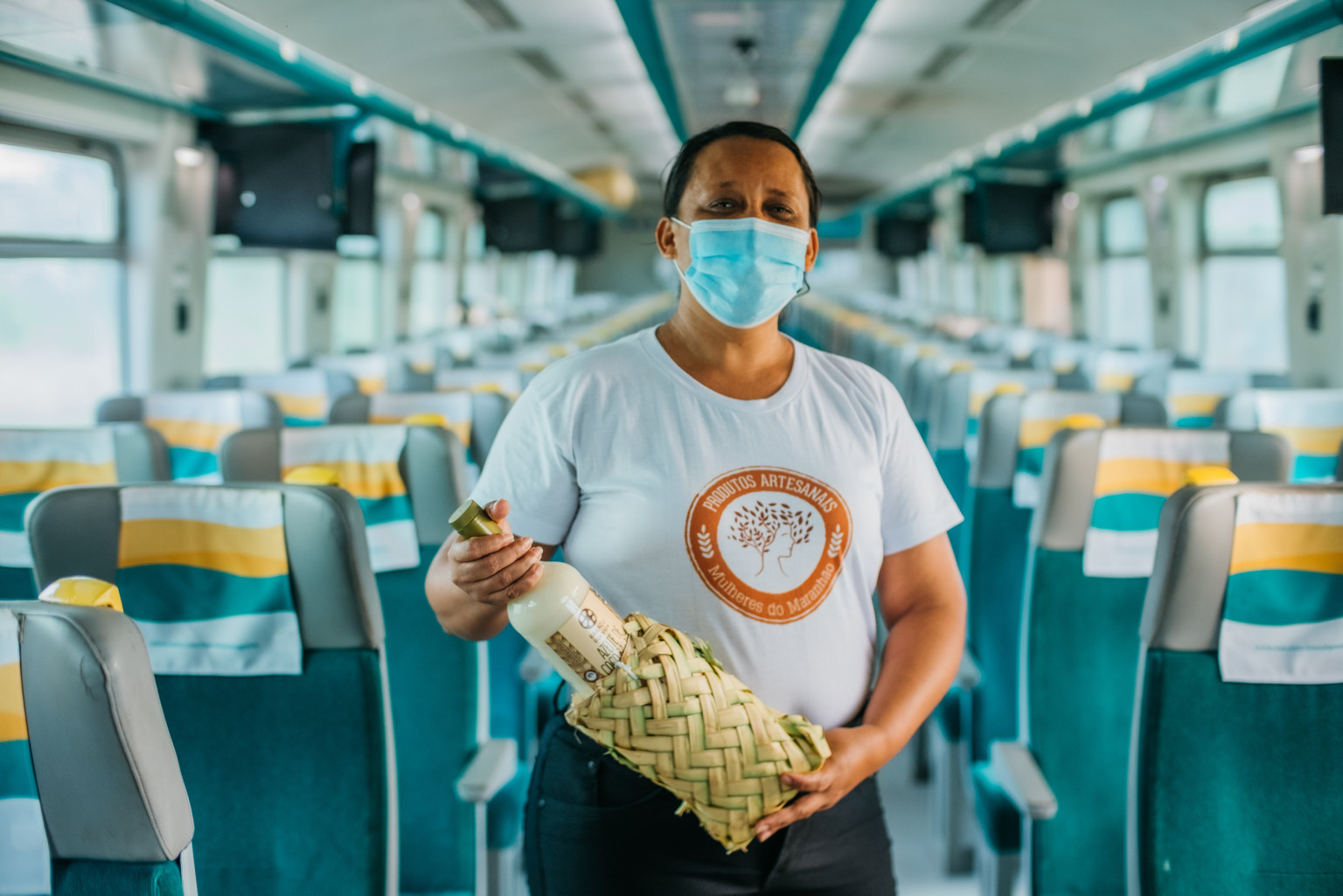 Photo of a participant from the Rede Mulheres do Maranhão (Women's Network of Maranhão) wearing a protective mask with the project's blouse securing a bottle of their product.