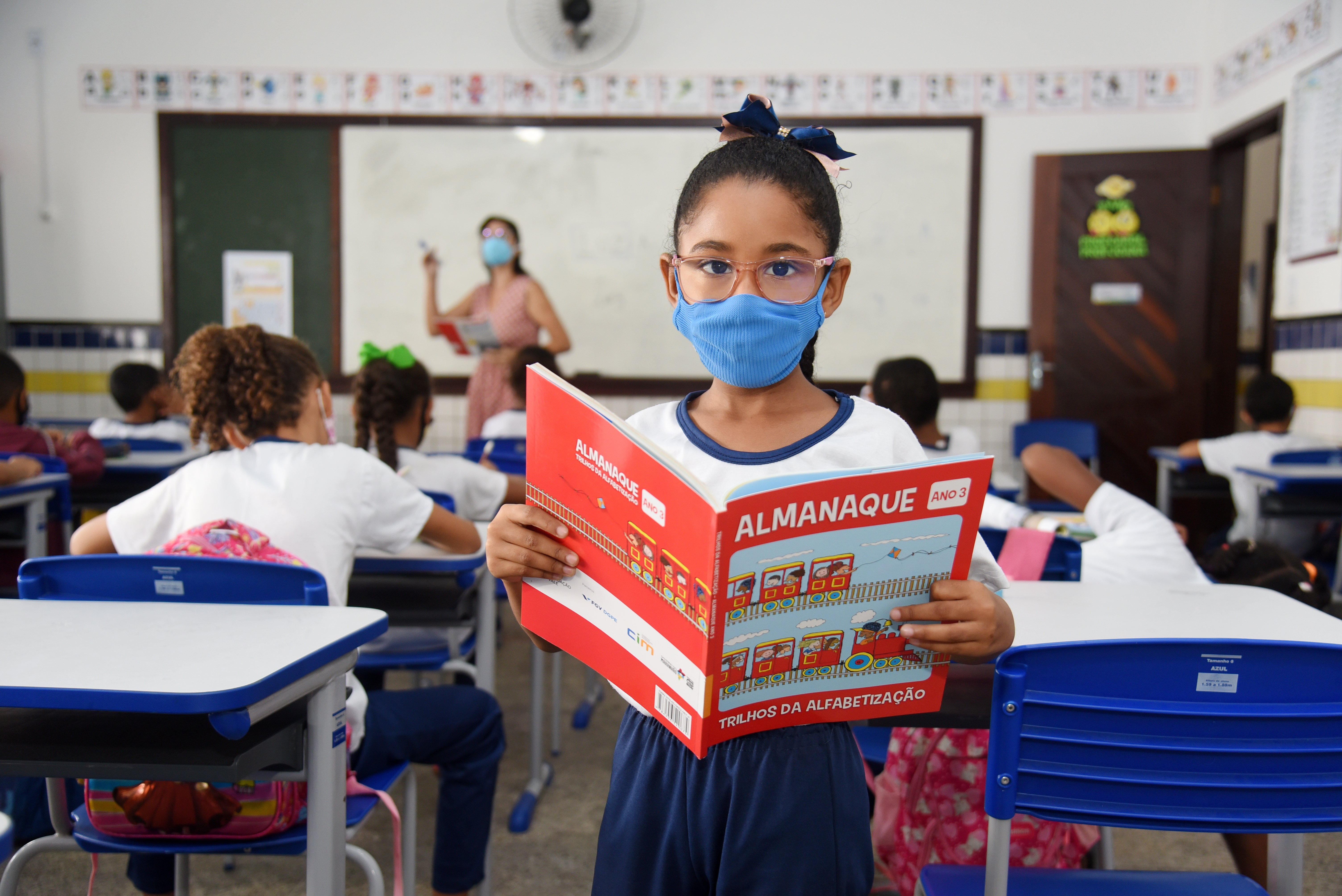 Image of a student who is part of the Vale Foundation's social initiatives. She is standing, wearing glasses and a mask, and holding a book. Behind her you can see the class with their attention focused on the teacher who is standing in front of the blackboard.