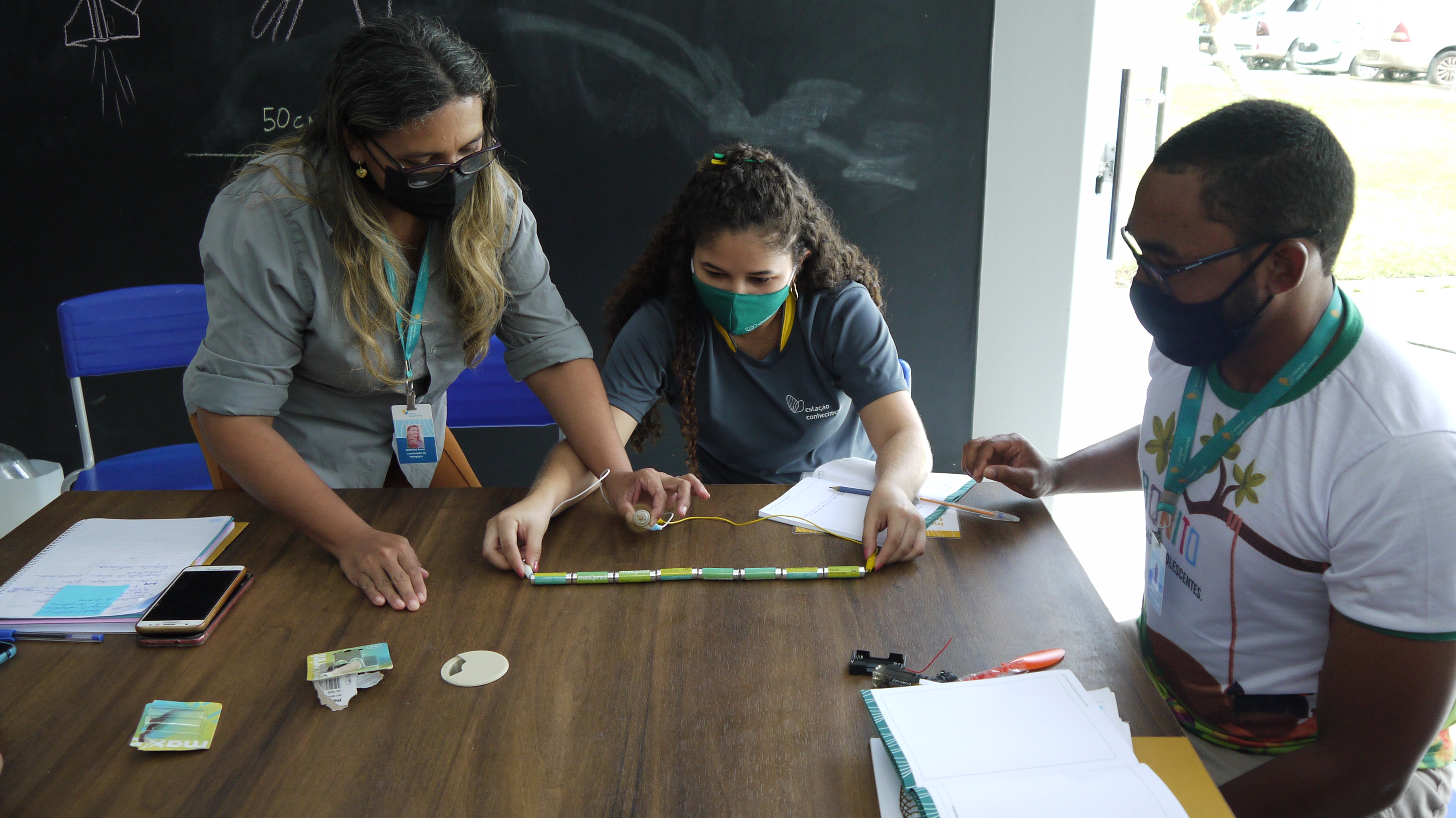 Imagem de três profissionais da área de educação científica e tecnológica reunidos em volta de uma mesa. Todos usam máscara. Eles estão com a atenção voltada para o teste do funcionamento de um motor, que é um objeto linear.