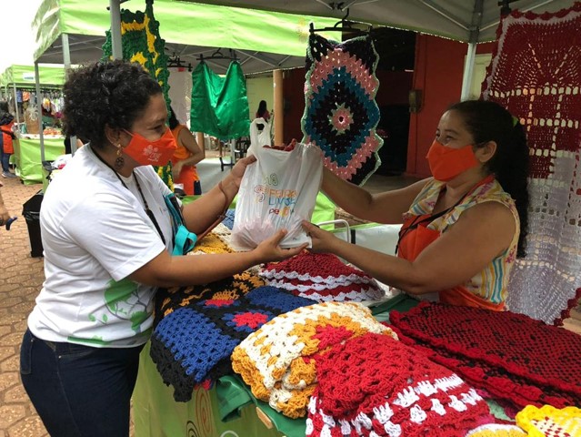 Image of two women, one a vendor and the other a customer, at the Feira Livre do Pequiá. They stand at a crochet stall and hold a bag.