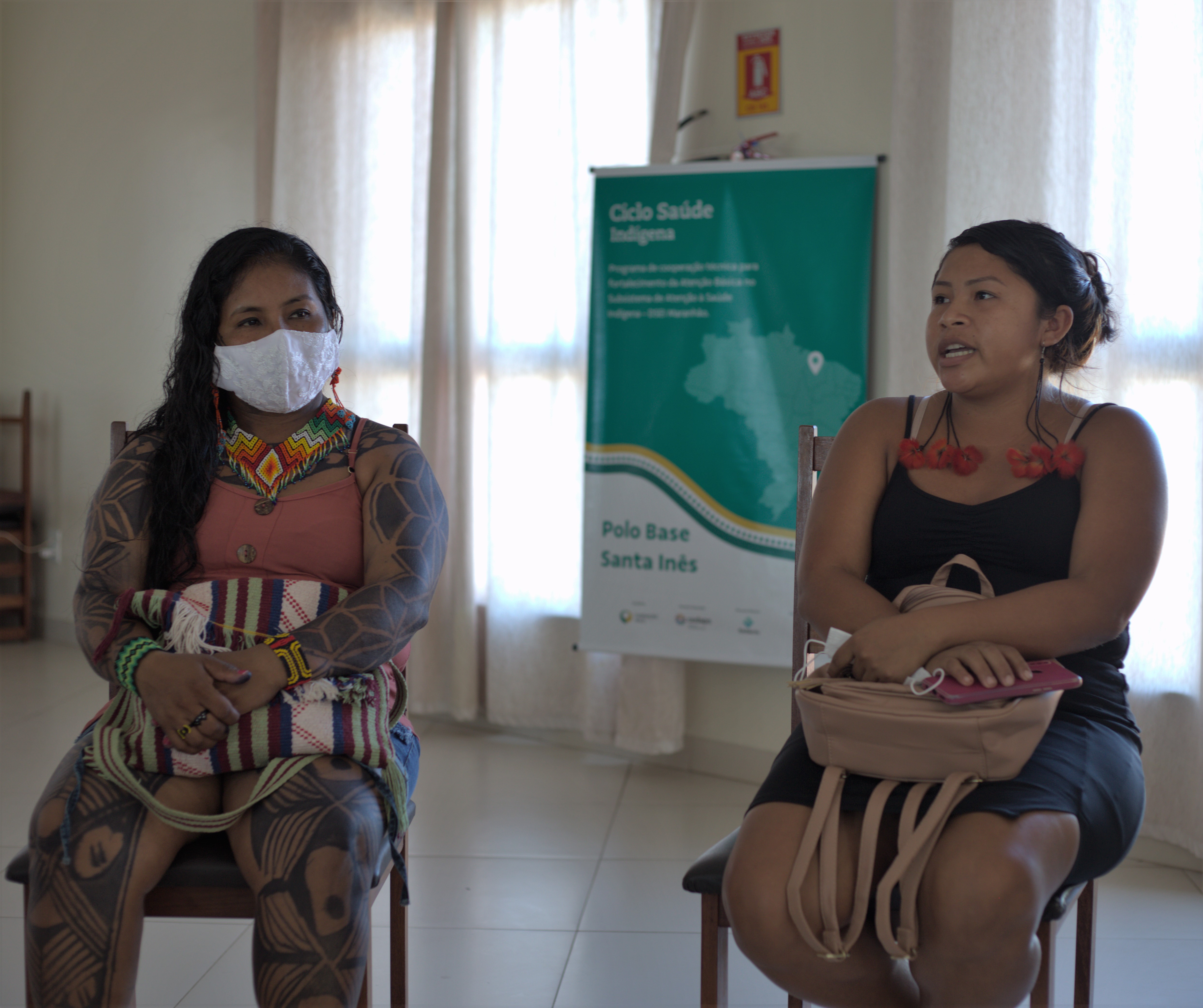 Imagem de duas mulheres indígenas sentadas em uma sala. Atrás delas é possível ver um cartas sobre o Ciclo de Saúde Indígena
