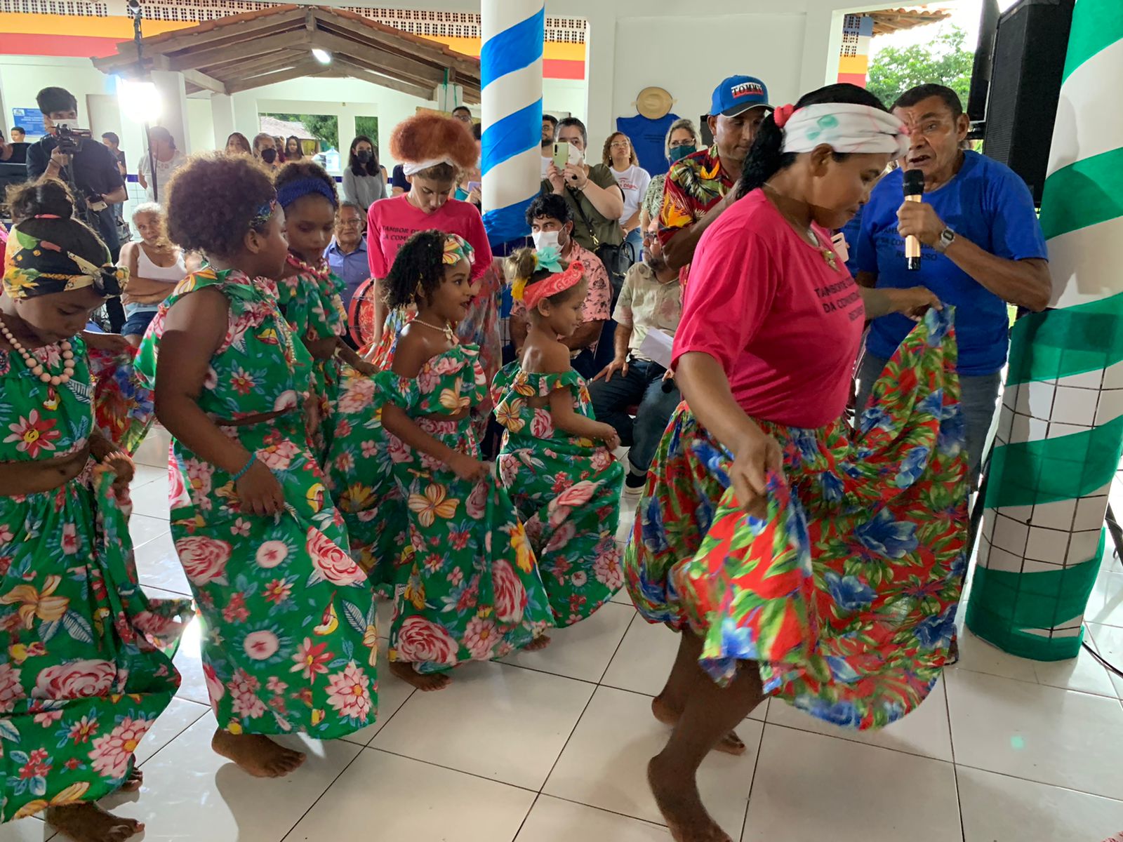 Conjunto de pessoas reunidas em evento que valoriza cultura quilombola. Na foto, mulheres e crianças dançam e usam vestidos verdes floridos e adereços na cabeça. Ao fundo pessoas fotografam.