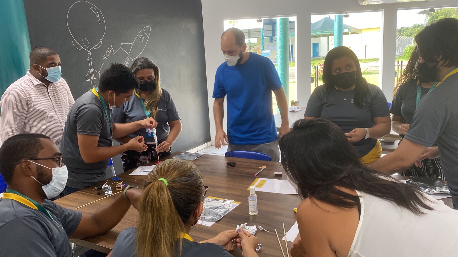 Imagem de diversas pessoas reunidas em volta de uma mesa, todas usam máscara e estão realizando trabalhos manuais.