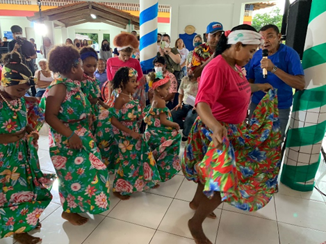 A group of people gathered in an event that values the quilombola culture. In the photo, women and children dance and wear green flowered dresses and head ornaments. In the background people taking pictures.