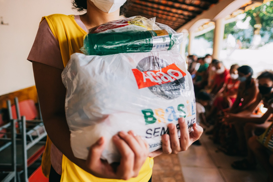 Imagem de uma pessoa segurando uma sacola com alimentos. Atrás dela é possível ver diversas pessoas sentadas. Todas usam máscaras de proteção.